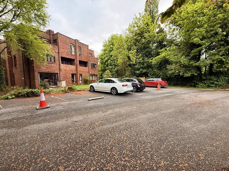 The car park beside the apartment block, edged by tall trees