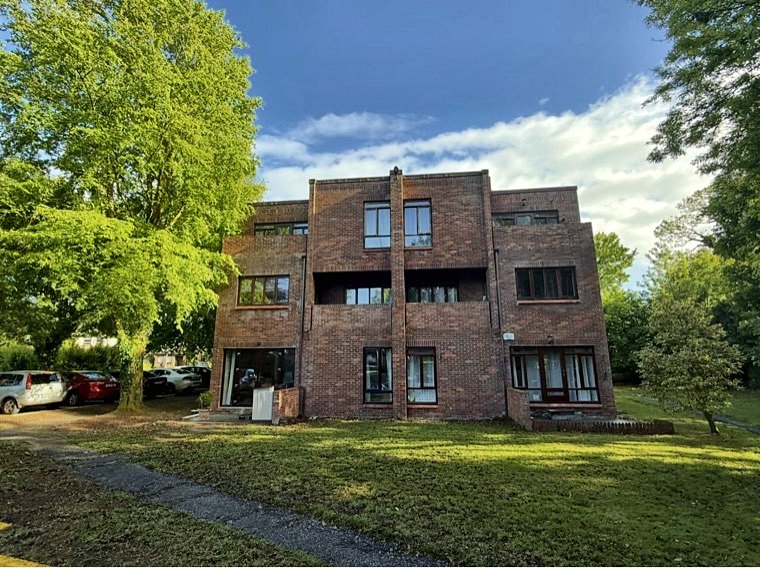 The front of the apartment block seen straight on across the lawn, with cars parked to the left