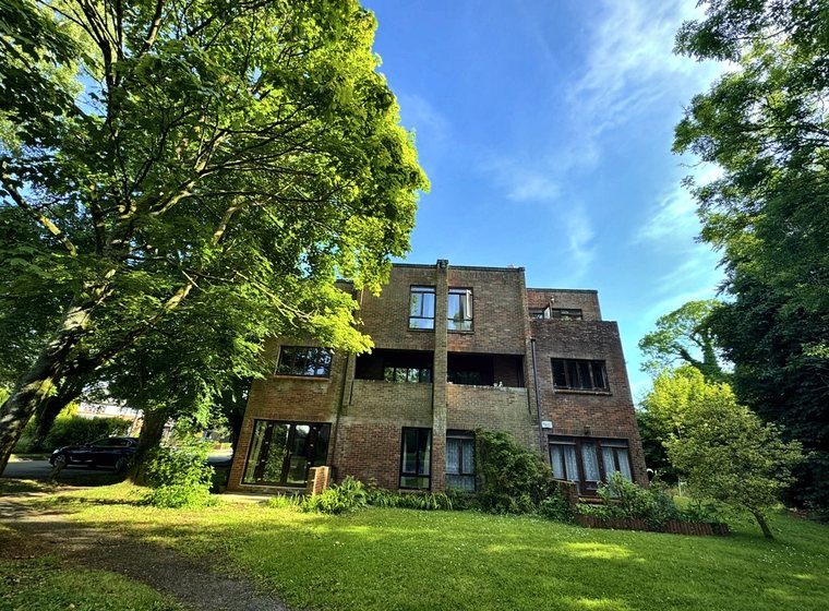 The apartment block seen across the lawn, framed by large trees in full leaf under a blue sky